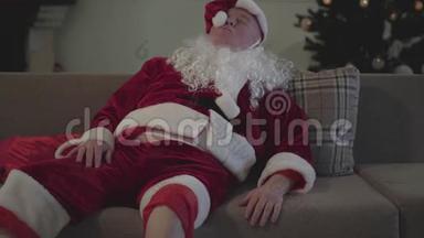 醉汉假白胡子穿着圣诞老人的服装躺在家里的沙发上，挥舞着手。 坏圣诞老人